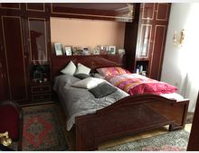 Schlafzimmer mit dunklen Holzmöbeln und eingebautem Schrank, der den gesamten hinteren Bereich einnimmt. Zwei Betten stehen nebeneinander, bedeckt mit grauer und bunter Bettwäsche. Auf dem Schrank stehen eingerahmte Fotos, und der Boden ist mit orientalen Teppichen ausgelegt.