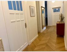 Flur in einer Wohnung mit hellem Holzparkett im Fischgrätmuster, weißen Türen mit Glaseinsätzen, einem großen Wandspiegel mit goldener Verzierung und einer roten Kommode mit Dekoration. Die Wände sind weiß und schlicht gestaltet.