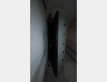 Ein schmaler, schlecht beleuchteter Gang mit Betonwänden, in dem eine massive Stahltür leicht geöffnet ist. Die Tür besitzt seitliche Verschlussbolzen, die typisch für einen Schutz- oder Tresorraum sind.