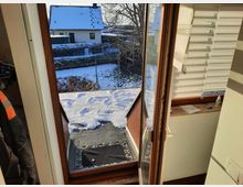 Blick durch eine geöffnete Tür ins Freie, mit einem schneebedeckten Garten sowie einem Haus mit verschneitem Dach im Hintergrund. Im Innenraum sind ein Fenstersims mit Gegenständen und ein Rollladen zu sehen.