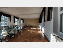 Großer, heller Raum mit Holzboden im Fischgrätmuster, weißen Wänden und Fenstern mit grünen Vorhängen. Der Raum ist mit Stühlen und Tischen ausgestattet, die an einer Seite gestapelt sind, und hat einen gelben Wandabschnitt am Kopfende.