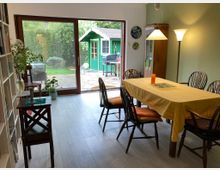 Esszimmer mit Holzboden und einem Holztisch, der mit einer gelben Tischdecke bedeckt ist, umgeben von Stühlen. Im Hintergrund führen eine große Glastür und Fenster zu einem Garten mit einer kleinen grünen Gartenhütte.