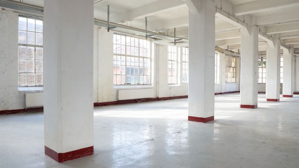 Großer, leerer Industrie- oder Loftinnenraum mit weißen Stützpfeilern, freiliegenden Deckenbalken und weitem Betonboden. Entlang der Außenwand befinden sich große, mehrteilige Sprossenfenster, die viel Tageslicht in den Raum lassen.