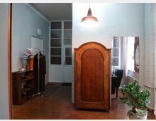 Wohnungsflur mit Holzboden, einem großen, antiken Holzschrank in der Mitte und einer hängenden Deckenleuchte. Im Hintergrund ist eine weiße Tür mit Glasfenstern sowie ein kleiner Kleiderständer und ein Regal mit Blumen zu sehen; rechts stehen eine Zimmerpflanze und ein runder Spiegel an der Wand.