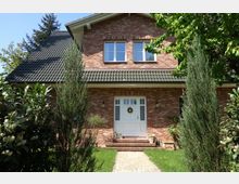 Backsteinhaus mit dunkelgrauem Satteldach, einer weißen Eingangstür mit Dekoration und zwei Fensterreihen. Der Vorgarten ist von grünen Büschen, Bäumen und einem gepflasterten Weg umgeben.