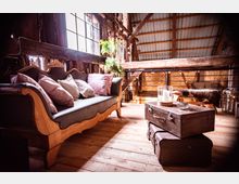 Gemütlicher Wohnbereich in einem rustikalen Dachgeschoss mit Holzboden, einer antik gestalteten Couch mit bunten Kissen, einem Fenster mit Holzrahmen und einem kleinen Tisch aus gestapelten Vintage-Koffern mit einer Kerze darauf. Im Hintergrund sind Holzbalken und Metallgeländer sichtbar.