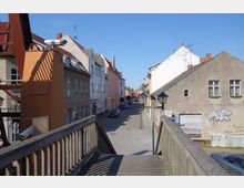 Straßenszene in einer kleinen Stadt, aufgenommen von einer Holzbrücke aus. Links und rechts der gepflasterten Straße stehen einfache Häuser mit teils renovierten, teils verfallenen Fassaden; im Vordergrund sind Straßenlaternen und ein orangefarbenes Metallgerüst sichtbar.