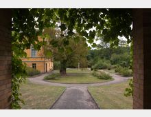 Blick durch einen von Weinlaub umrahmten Torbogen auf einen historischen Garten mit geschwungenen Wegen, Rasenflächen und Büschen, im Hintergrund ein gelbes Gebäude mit großen Fenstern und ein Baum.