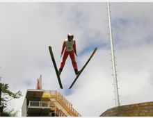 Ein Skispringer in rotem Anzug und Helm ist während eines Sprungs in der Luft zu sehen, mit Skiern in V-Position. Im Hintergrund befinden sich eine hölzerne Skisprungschanze, ein Metallmast und ein bewölkter Himmel.