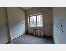 Ein leerer, verfallener Raum mit abblätternder Farbe an den Wänden, einem rostigen Heizkörper unter einem vergitterten Fenster und sichtbaren Rohren in der Ecke. Durch das Fenster ist ein Außenbereich mit Backsteinmauern und kahlen Bäumen zu sehen.