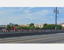 Straßenbrücke mit Straßenbahnschienen und einem Geländer mit rot-weißen Baustellenmarkierungen. Im Hintergrund sind Bäume und mehrstöckige Wohngebäude sowie einzelne moderne Hochhäuser sichtbar, unter einem bewölkten, blauen Himmel.