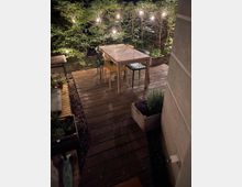 Eine Terrasse aus Holz mit einem rechteckigen Esstisch und unterschiedlich farbigen Stühlen, umgeben von Pflanzen. Im Hintergrund stehen Büsche, die mit Lichterketten beleuchtet sind, und Blumenkästen befinden sich am Rand der Terrasse.