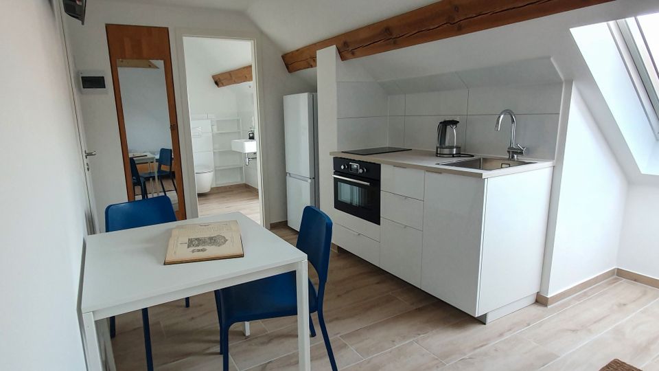 Heller Dachgeschossraum mit kleiner Küchenzeile in Weiß, integriertem Backofen, Kühlschrank und Spüle unter einer Dachschräge mit Dachfenster. Im Vordergrund steht ein weißer Esstisch mit blauen Stühlen; sichtbare Holzbalken und eine Türöffnung führen in ein angrenzendes Bad.