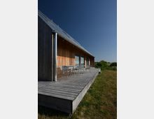 Ein modernes Holzhaus mit geneigtem Dach und Metallverkleidung an der Dachkante, umgeben von einer Terrasse aus Holzdielen. Auf der Terrasse stehen ein Glastisch und Metallstühle, die auf eine Wiese mit weiter Landschaft im Hintergrund blicken.