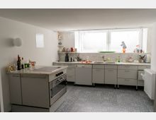 Helle Küche mit weißen Wänden und grauer Einrichtung, bestehend aus einem Herd mit Marmorkochplatte und mehreren Schränken sowie einer Arbeitsfläche mit Spüle. Ein großes Fenster lässt Tageslicht herein und der Boden ist mit blau-gemusterten Fliesen ausgelegt.