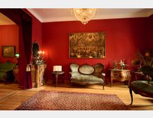 Wohnzimmer im historischen Stil mit roten Wänden, antiken Möbeln und einem Kristall-Kronleuchter. Zentrales Element ist ein grünes Sofa vor einer Wand mit einem Gemälde, flankiert von dekorativen Kommoden und einem Teppich auf Holzboden.