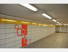 Unterführung mit hellen Fliesenwänden, die teilweise ein gelbes Band oben und ein rotes Muster aus Kreisen auf der Seite zeigen. Der Boden ist aus grauem Stein, und die Decke ist mit langen, rechteckigen Leuchtstofflampen ausgestattet.