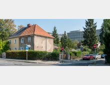 Ecke einer Wohnstraße mit einem zweistöckigen Haus mit rotem Ziegeldach, umgeben von Bäumen und Hecken. Im Hintergrund sind ein modernes Gebäude und mehrere parkende Autos zu sehen; Straßenschilder, darunter ein Taxistand- und ein Einfahrt-verboten-Schild, befinden sich im Vordergrund.