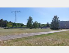 Offene Grünfläche mit Rasen, durchzogen von einem schmalen Gehweg. Im Hintergrund stehen einzelne Bäume, ein Strommast, ein langes Flachgebäude und ein mehrstöckiges Wohnhaus, umgeben von weiteren Bäumen und Wald.