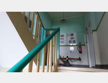 Treppenhaus mit hellgrünen Wänden und einem Holzgeländer mit türkisfarbener Handlaufleiste. An der Wand sind gerahmte Bilder aufgehängt, daneben ein Feuerlöscher; der Boden ist mit einem hellen Belag versehen.