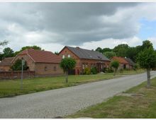 Eine ländliche Straße mit Kopfsteinpflaster, gesäumt von niedrigen roten Backsteinhäusern mit Satteldächern und kleinen Vorgärten. Im Hintergrund sind Bäume zu sehen, und der Himmel ist teilweise bewölkt.