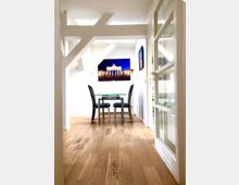 Heller, schmaler Innenraum mit sichtbaren weißen Dachbalken und hellem Holzboden, der zu einer kleinen Essecke führt. Am Ende steht ein Glastisch mit zwei schwarzen Stühlen, darüber hängt ein Wandbild; rechts sind große verglaste Tür- oder Fensterrahmen.