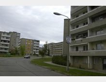 Straßenszene in einem Wohngebiet mit mehreren Plattenbau-Wohnhäusern, umgeben von Grünflächen und Gehwegen; im Vordergrund verläuft eine asphaltierte Straße, und links parkt ein Auto.