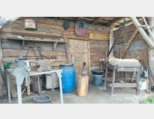 Holzschuppen mit Schmiedeausrüstung, darunter ein Amboss, Metallwerkzeuge, ein großer blauer Kunststoffbehälter und eine Werkbank. Die Wände bestehen aus grob gehauenen Holzbalken, und verschiedene handwerkliche Geräte hängen an den Wänden.