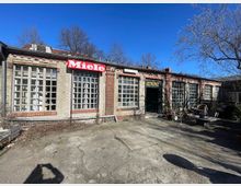 Flacher Industrie- oder Werkstattbau aus Ziegel und Beton mit großen Sprossenfenstern und einem roten „Miele“-Schild an der Fassade. Davor liegt ein asphaltierter Hof mit Bänken und kahlen Bäumen unter blauem Himmel.