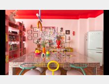 Bunter, moderner Essbereich mit auffälliger, mosaikbedeckter Tischplatte, umgeben von pastellrosa Wänden. Im Hintergrund stehen ein Retro-Kühlschrank, offene Regale mit Büchern und Dekorationsgegenständen sowie farbenfrohe Kunstwerke an der Wand.