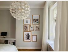 Helles Schlafzimmer oder Arbeitszimmer mit einer dekorativen weißen Hängeleuchte in Blumenoptik, mehreren gerahmten Bildern an einer Wand und einem Schreibtisch mit Computer in einer Ecke. Rechts befindet sich ein großes Fenster mit langen weißen Vorhängen.