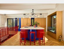 Moderne Küche mit buntem Design: Eine zentrale Kochinsel mit weißer Arbeitsfläche und roten, gerillten Elementen wird von drei roten Barhockern umgeben. Im Hintergrund befinden sich Holzverkleidungen, blaue Akzente, eine große Fensterfront sowie eine Sitznische mit Holzeinfassung und Kissen.