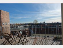 Dachterrasse mit Holzboden, einem kleinen Tisch und zwei Stühlen aus Holz, umgeben von einem Metallgeländer. Im Hintergrund sind Dächer, einige Bäume und Baukräne unter einem blauen, leicht bewölkten Himmel sichtbar.