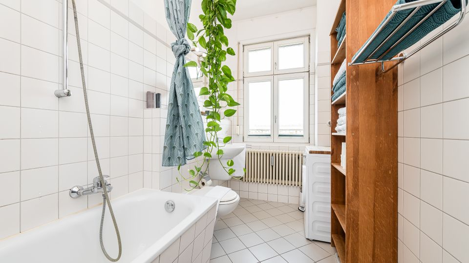 Helles, gefliestes Badezimmer mit Badewanne und Duscharmatur an der linken Wand sowie einem großen Fenster über einem Heizkörper am Ende des Raums. Rechts steht ein Holzregal mit Handtüchern neben einer Waschmaschine; eine hängende Grünpflanze und ein gemusterter Duschvorhang setzen Akzente.