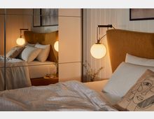 Schlafzimmer mit einem Bett, das ein senfgelbes Kopfteil hat, weiße Bettwäsche und ein Kissen mit abstraktem Gesichtsmuster. Daneben ist eine runde, leuchtende Wandlampe und ein Nachttisch mit einem Glas Wasser, einer Brille und einem dekorativen Zweig sichtbar. Im Hintergrund reflektieren Spiegel die Szene.