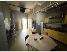 Schmaler Pausenraum mit Tisch und Stühlen im Vordergrund sowie einer Küchenzeile mit gelben Schränken, Spüle und offenen Regalen an der rechten Wand. Am Ende des Raums führen mehrere Türen in angrenzende Bereiche; an der linken Seite stehen ein Wasserspender und ein Servierwagen.