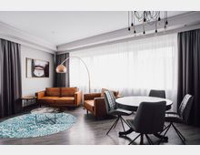 Modern eingerichtetes Wohnzimmer mit braunen Ledersofas, einem blauen Teppich mit Ornamenten, einem runden Glastisch und einer gebogenen Stehlampe mit kupferfarbenem Lampenschirm. Im Hintergrund bodentiefe Fenster mit weißen Gardinen und grauen Vorhängen, daneben ein Essbereich mit runden Tisch und gepolsterten schwarzen Stühlen.