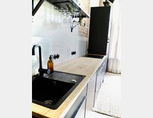 Moderne Küche mit einer langen Arbeitsplatte aus hellem Holz und schwarzen Unterschränken. Sichtbare Details umfassen ein schwarzes Spülbecken mit Armatur, über der Arbeitsplatte hängende Weingläser an einem Metallhalter und eine ausgekleidete Rückwand aus Betonoptik und teilweise freigelegtem Ziegelmauerwerk.