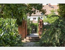 Eingang eines Gebäudes mit weißen Wänden und einem rot gedeckten Ziegeldach, eingerahmt von einer roten Backsteinmauer und zwei kleinen gemauerten Säulen. Der Weg führt durch üppige grüne Vegetation und endet an einer mit Laub überdachten Tür.