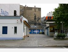 Straßenszene mit einer kleinen Tankstelle auf der rechten Seite und einem weiß gestrichenen Gebäude links; im Hintergrund eine alte Backsteinwand mit verblasster Fassade und Graffiti, sowie blaue Garagentore. Ein Auto ist in der Einfahrt geparkt, umgeben von Betonboden und Pflastersteinen.