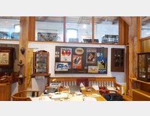 Ein Innenraum eines Museums oder Antiquitätengeschäfts mit Holzmöbeln, antiken Vitrinen, Werbetafeln und gerahmten Plakaten an der Wand. Im Vordergrund ein Schreibtisch mit Papieren und einem Laptop, im Hintergrund Holzsäulen und ein Obergeschoss mit weiteren Ausstellungsgegenständen.
