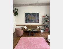 Wohnzimmer mit Holzboden und dekorativer Stuckdecke. Im Raum stehen ein braunes Sofa, ein rosa Sessel, ein grüner Couchtisch und ein rosa Teppich. An der Wand hängt eine Wanddekoration mit Pflanzen- und Figurenmotiven, daneben ein Regal mit Büchern und Dekorationsobjekten.