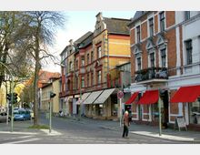 Straßenszene mit historischen Gebäuden aus Klinker und Putz in rot-gelben Farbtönen, darunter mehrere Geschäfte mit roten Markisen. Im Vordergrund befinden sich eine Straße mit Fußgängerüberweg, Verkehrsampeln sowie ein älterer Mann, der die Straße überquert.