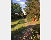 Ein grüner Garten mit einem gepflasterten Weg, umgeben von Bäumen, Sträuchern und Blumenbeeten. Im Hintergrund stehen eine große Tanne, eine rote Holzbank und dekorative Laternen.