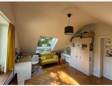 Dachgeschosszimmer mit schräger Decke und großem Dachfenster, das Tageslicht hereinlässt. Der Raum enthält einen gelben Zweisitzer, einen Schreibtisch mit weißem Stuhl, einen weißen Kleiderschrank und ein Sideboard mit Spiegel; der Boden ist aus Holzparkett.