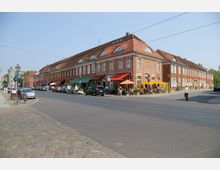 Straßenszene mit zweigeschossigen Backsteingebäuden und roten Ziegeldächern; im Erdgeschoss befinden sich Cafés mit bunten Markisen und Sonnenschirmen. Im Vordergrund eine gepflasterte Straße mit parkenden Autos und Fahrrädern, im Hintergrund ein historisches Tor.