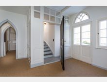 Heller Eingangsbereich eines Hauses mit weiß gestrichenen Wänden, einer schwarzen Eingangstür mit goldfarbenem Türgriff, großen Fenstern mit Sprossen und einem halbkreisförmigen Oberlicht. Eine Treppe mit grauen Stufen führt nach oben, und ein gewölbter Durchgang verbindet diesen Raum mit dem dahinterliegenden Flur.