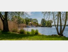 Grüne Uferlandschaft mit Bäumen und Schilf am Rand eines ruhigen Sees, im Hintergrund kleinere Boote an einem Steg sowie ein Haus, umgeben von Bäumen. Der Himmel ist klar und sonnig.