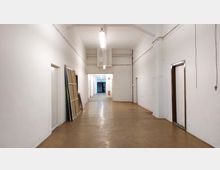 Langer, schlichter Flur mit weißen Wänden und braunem Bodenbelag, an den Seiten Türen und an der linken Wand mehrere lehnende Rahmen. Am Ende des Flurs ist ein Durchgang in einen weiteren, beleuchteten Bereich zu sehen.