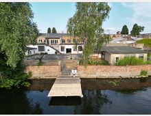 Ehemaliges Industriegebäude mit Backsteinfassade und Flachdachanbauten, umgeben von einer niedrigen Ziegelmauer. Im Vordergrund ein hölzerner Steg am Ufer eines Flusses mit Wasserpflanzen und einem Baum auf der linken Seite.